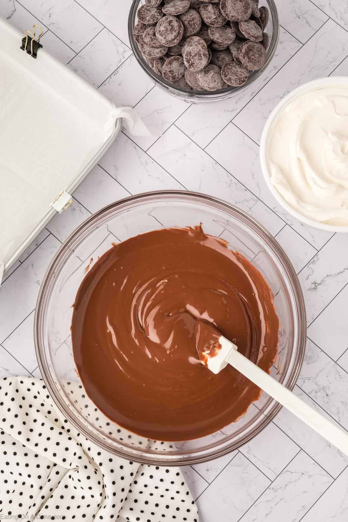 A bowl of melted chocolate with a spatula, next to a bowl of chocolate chips, Cool Whip, a baking pan lined with parchment paper, and a polka dot cloth on a tiled surfaceโperfect for making sweet homemade candy.