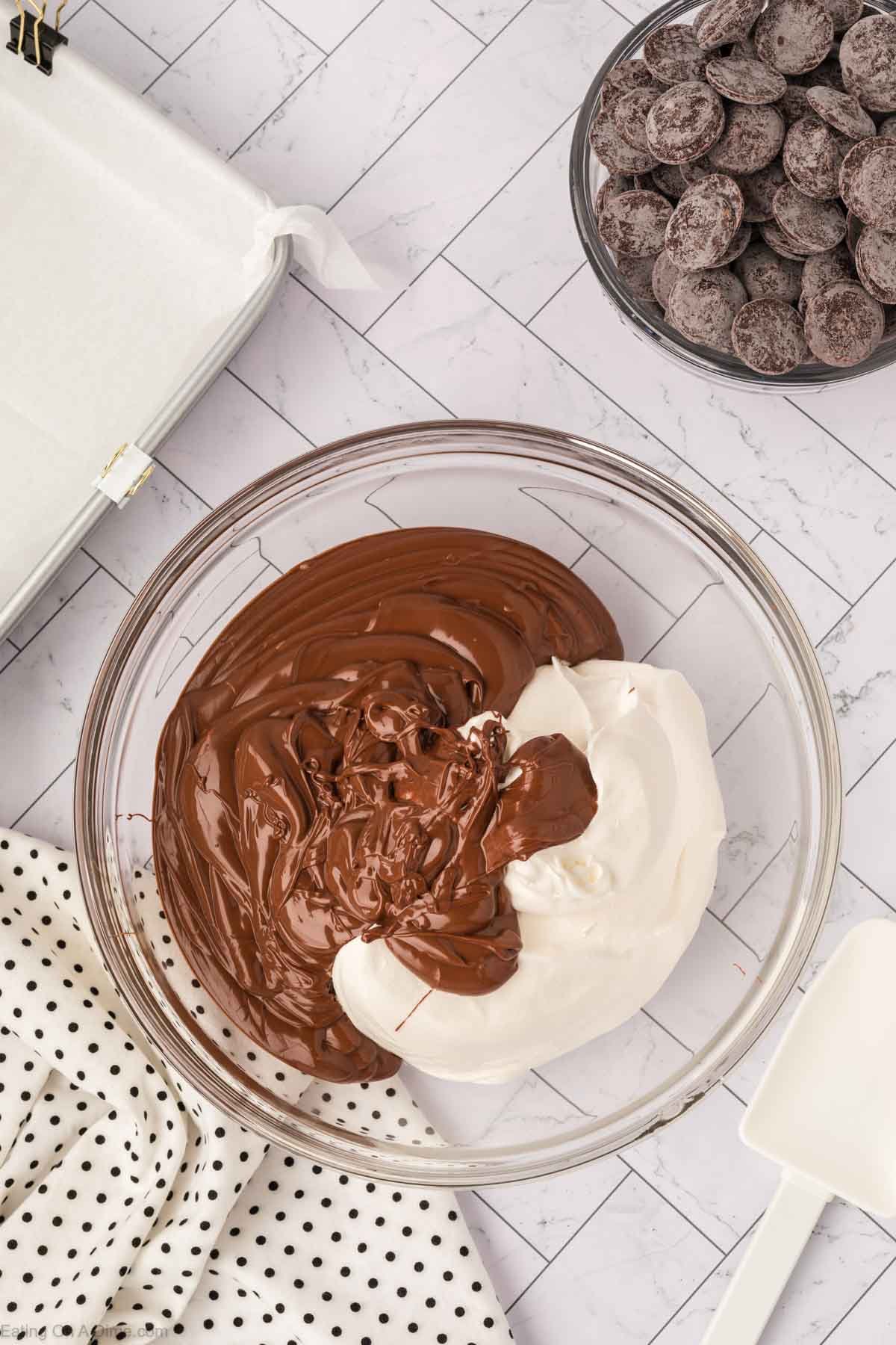 A glass bowl with melted chocolate and marshmallow fluff sits on a marble countertop, alongside Candy wafers, a white spatula, a polka dot cloth, and a parchment-lined baking pan.