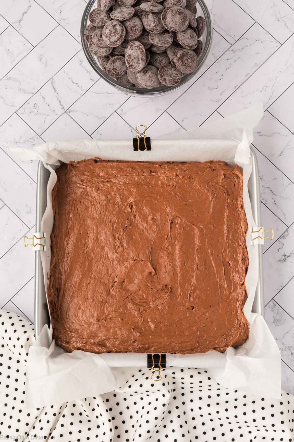 A square baking pan lined with parchment paper and filled with chocolate batter, secured at the edges with binder clips. Candy pieces and a bowl of chocolate sit nearby, along with a white cloth with black polka dots on a marble surface.