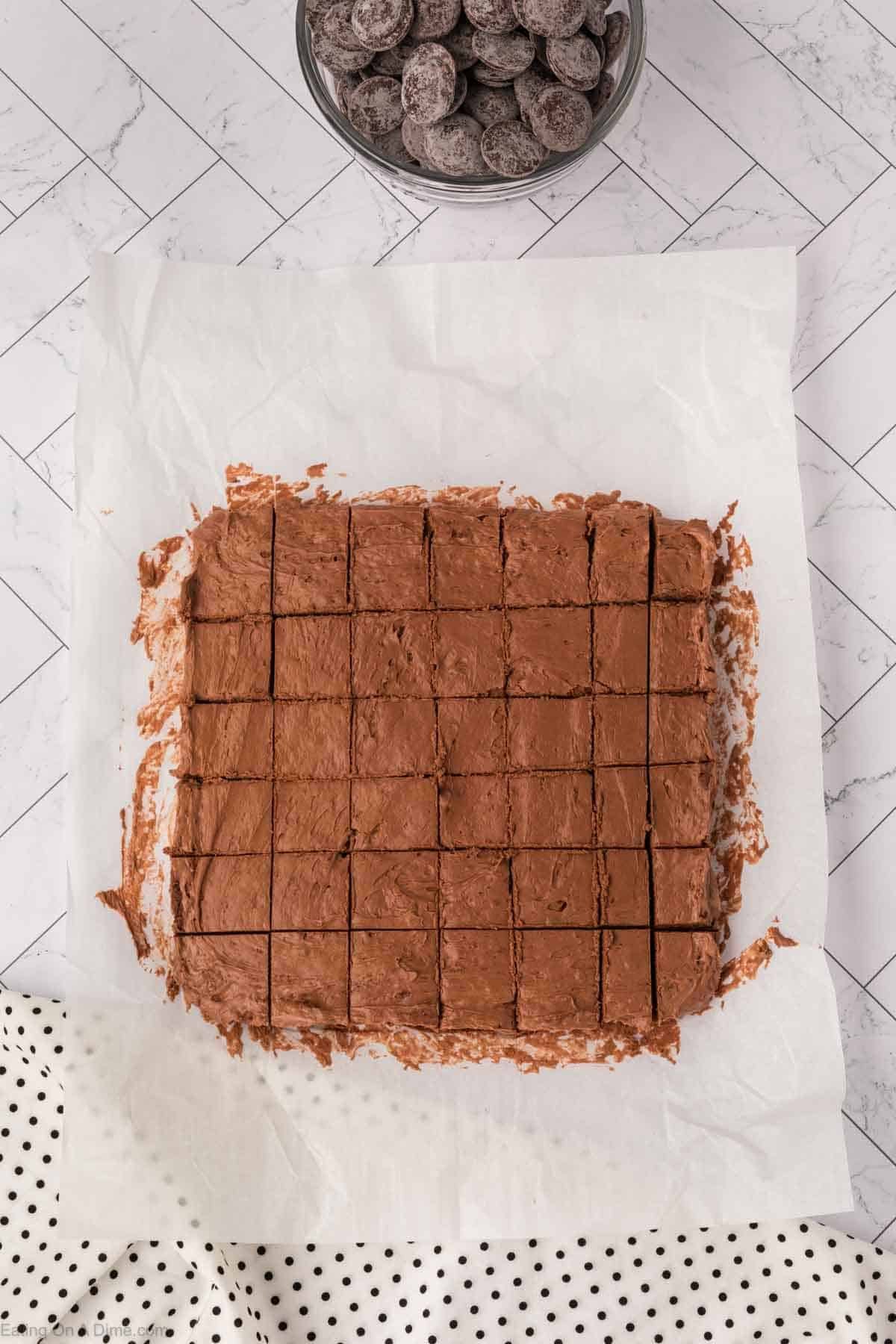 A square pan of Cool Whip Candy chocolate fudge, cut into neat squares, sits on parchment paper atop a white countertop. A bowl of chocolate pieces is at the top, and a white polka-dot cloth rests in the bottom corner.