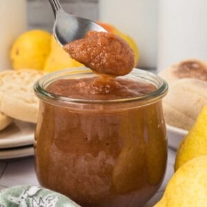 A spoon lifts a dollop of thick pear butter from a glass jar, surrounded by crumpets and fresh pears—an inviting scene that hints at a delicious pear butter recipe ready to be enjoyed.