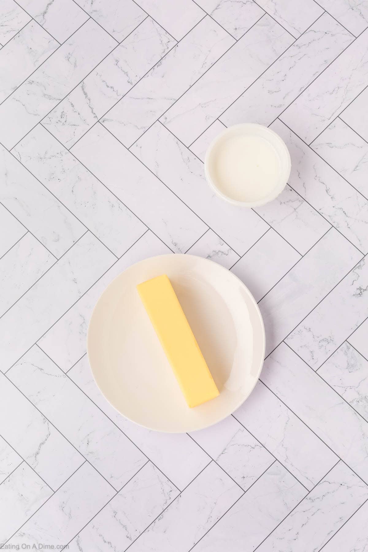 A stick of whipped butter on a white plate and a small bowl of milk rest on a white tile surface with a herringbone pattern.
