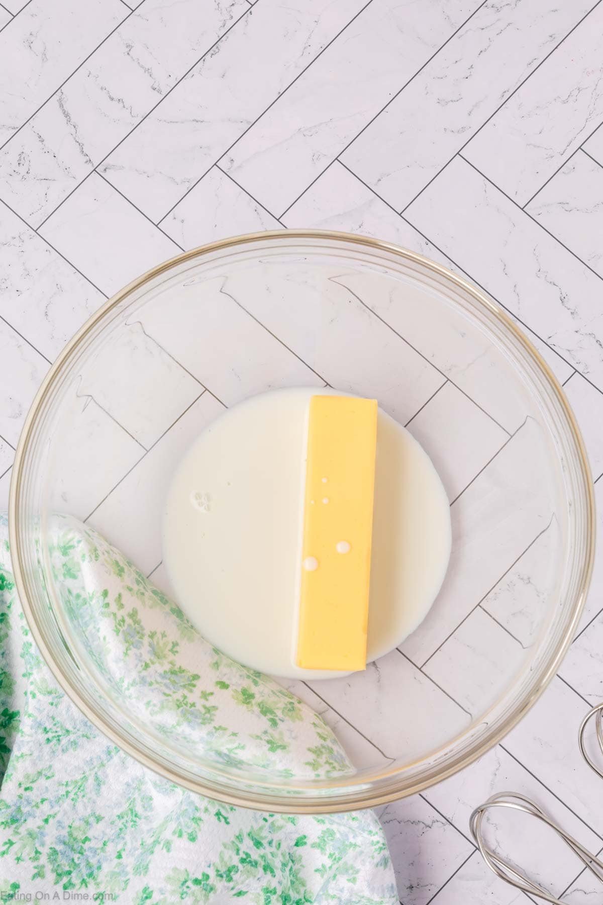 A glass bowl containing whipped butter and some milk sits on a white tiled surface, next to a green patterned towel and metal beaters.