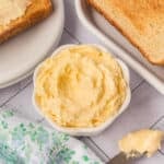 A small bowl of creamy whipped butter sits on a table beside a butter knife, slices of toasted bread, and a floral-patterned cloth.