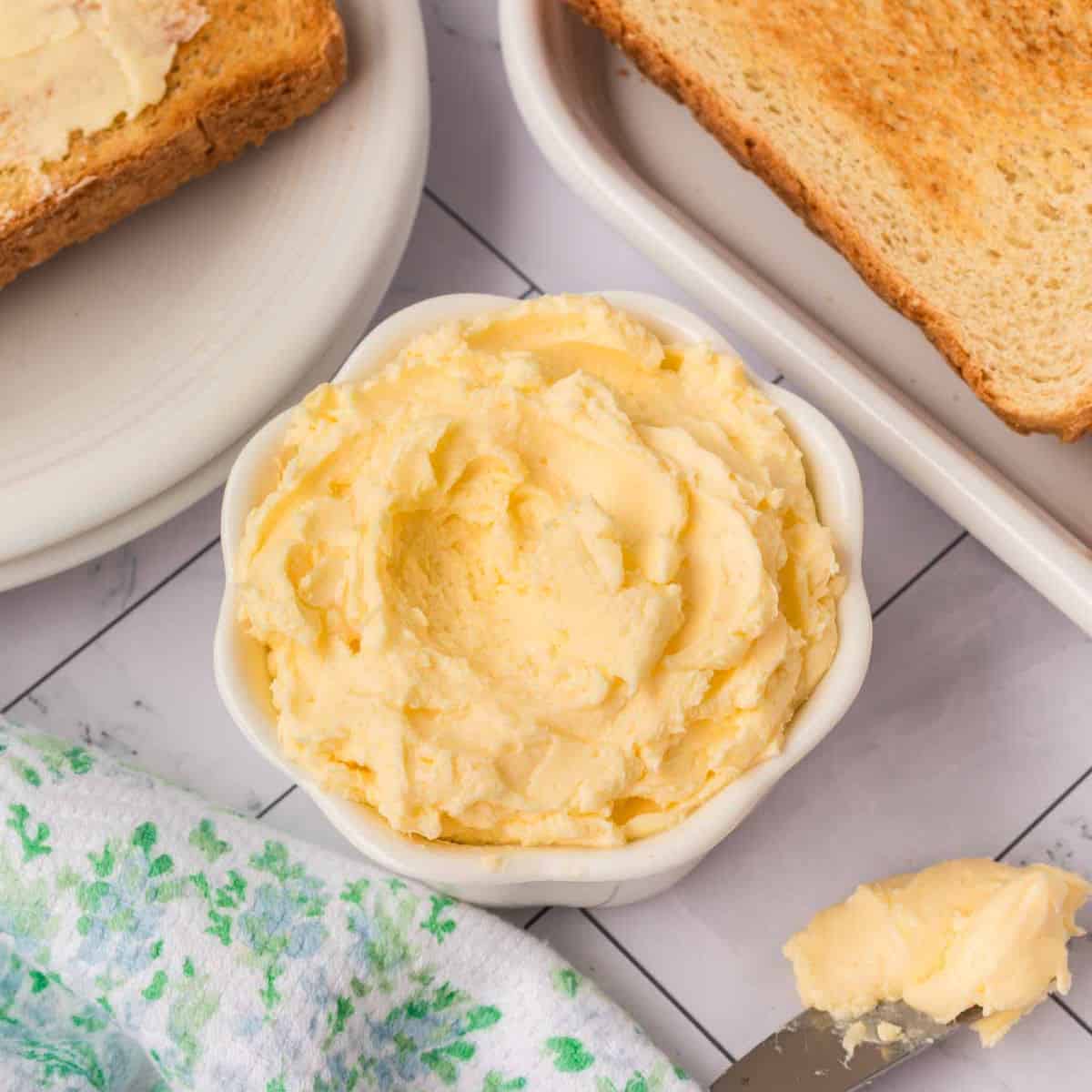 A small bowl of creamy whipped butter sits on a table beside a butter knife, slices of toasted bread, and a floral-patterned cloth.