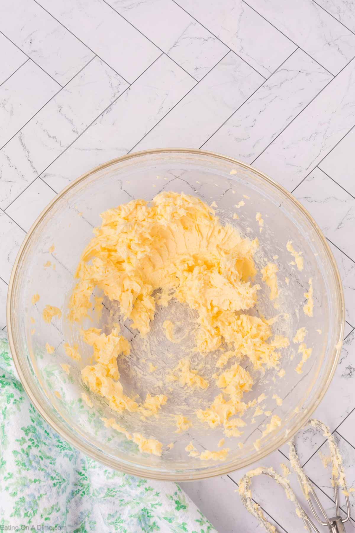 A glass bowl of whipped butter rests on a white tiled surface beside a green and white cloth and an electric hand mixer with some whipped butter clinging to the beaters.