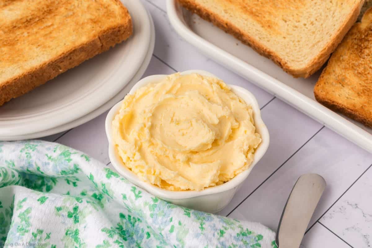 A small white bowl filled with creamy whipped butter sits on a white tiled surface, surrounded by slices of toasted bread on plates and a green-patterned cloth nearby. A butter knife is also visible.