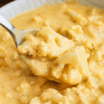 A close-up of a spoon scooping creamy, cheesy Cauliflower Mac and Cheese from a bowl of rich, yellow cheese sauce, with chunks of cauliflower visible throughout the dish.