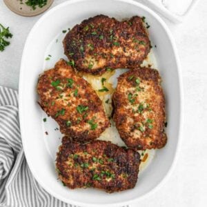 Four golden-brown, seasoned baked chicken breasts garnished with chopped parsley are arranged in a white baking dish on a light surface. Perfect for Meal Plan 88, a striped towel and a small dish of herbs are nearby.