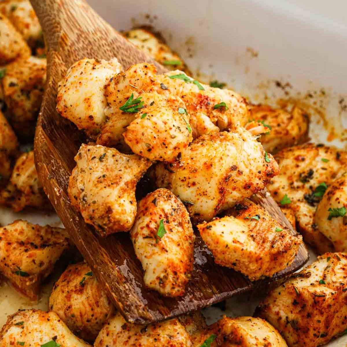 A wooden spoon scoops up Baked Chicken Bites, seasoned and garnished with chopped herbs, from a white dish. The golden brown chicken pieces are coated in visible spices and have a slightly crispy texture.