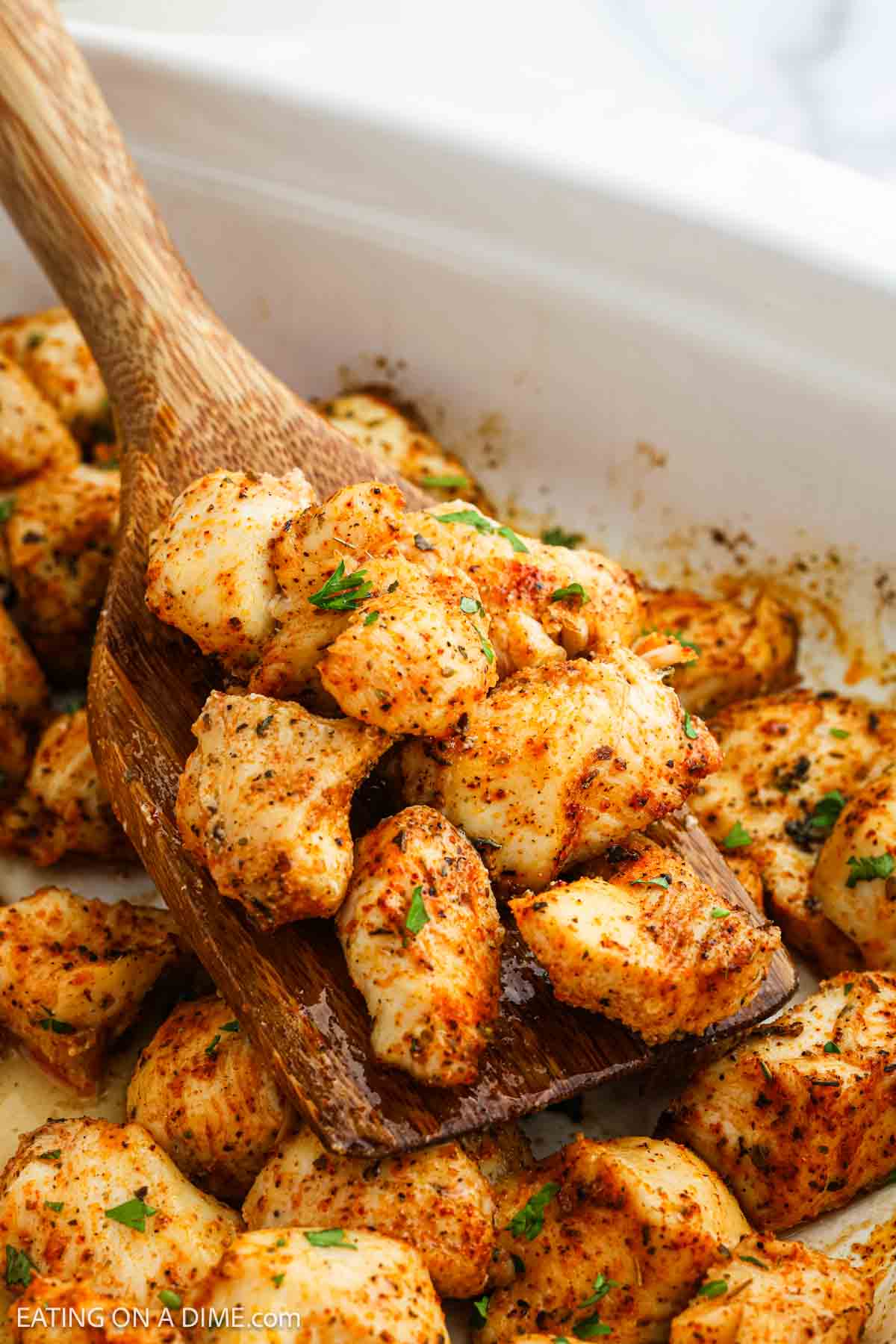 A wooden spoon scoops up seasoned Baked Chicken Bites from a white dish. The chicken is golden brown and garnished with chopped herbs.