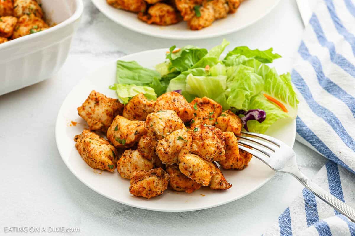 A white plate with seasoned Baked Chicken Bites and a side of fresh green salad. A fork rests on the plate, with a blue and white striped napkin nearby. Another dish with more chicken bites is partially visible in the background.