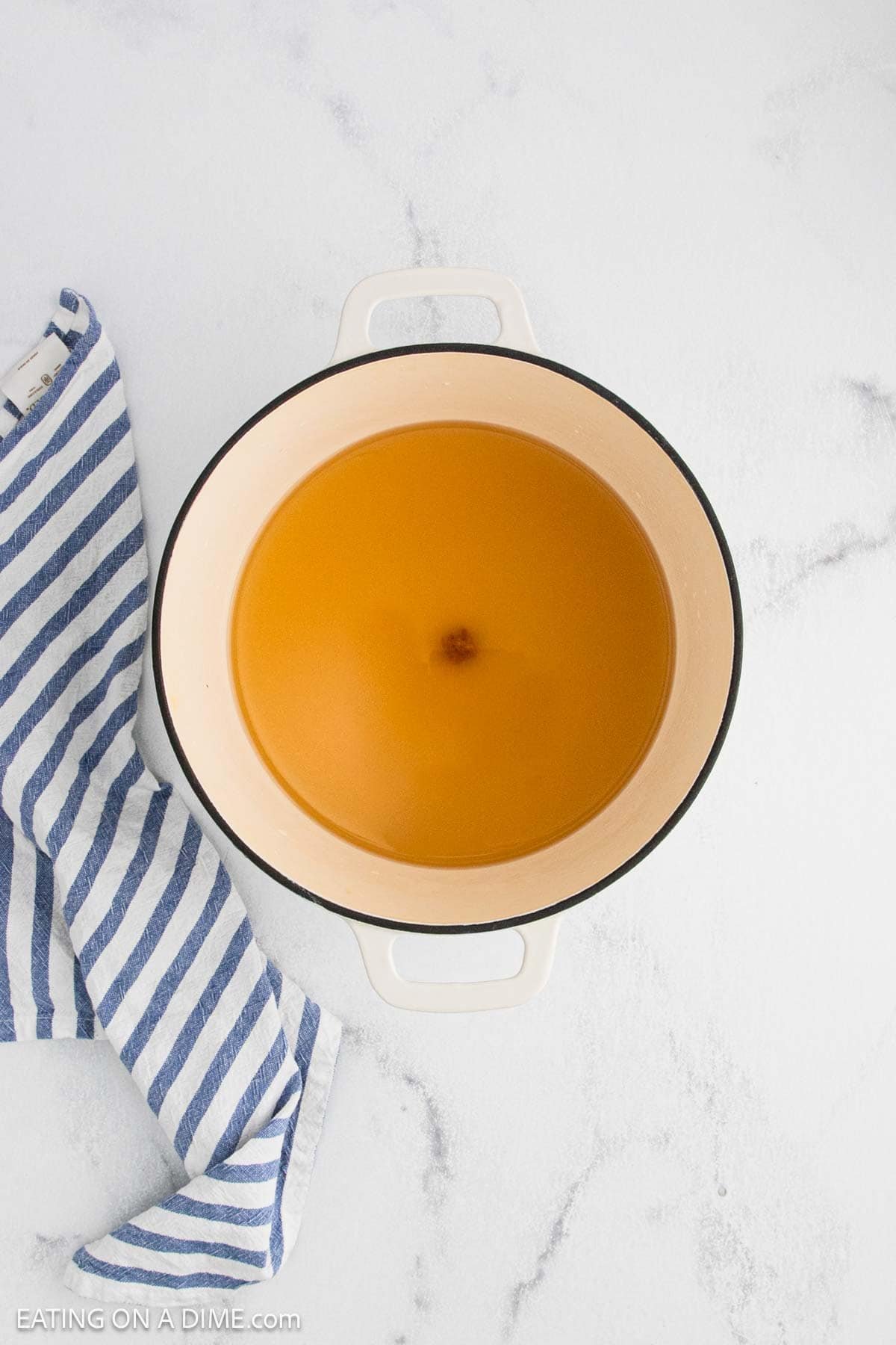 A white pot filled with golden brown broth and tender noodles sits on a white marble surface next to a blue and white striped kitchen towel, reminiscent of a cozy cafeteria meal.