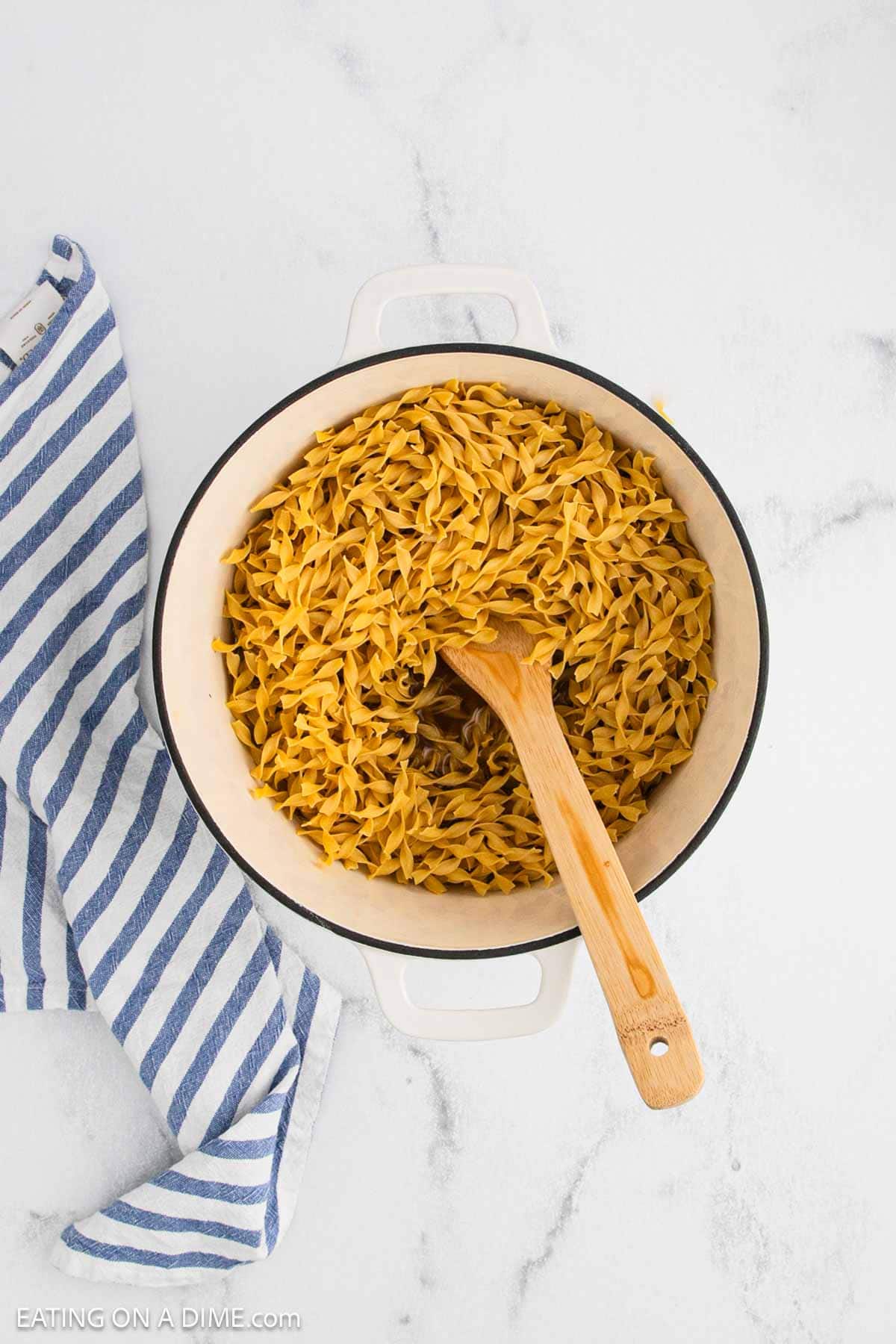 A white pot filled with cooked cafeteria noodles sits on a white surface, with a wooden spoon resting inside. A blue and white striped kitchen towel is draped nearby.