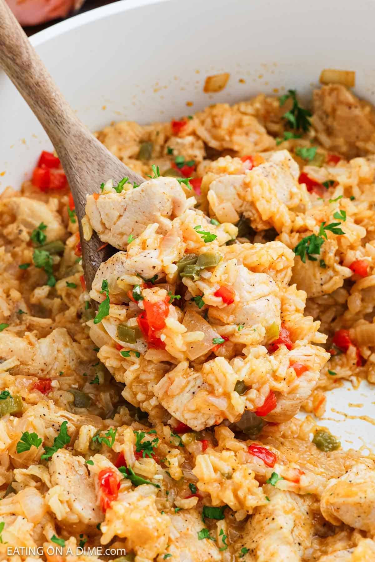 A wooden spoon scooping a serving of Cajun Chicken and rice with diced red and green bell peppers from a pan, garnished with chopped parsley.