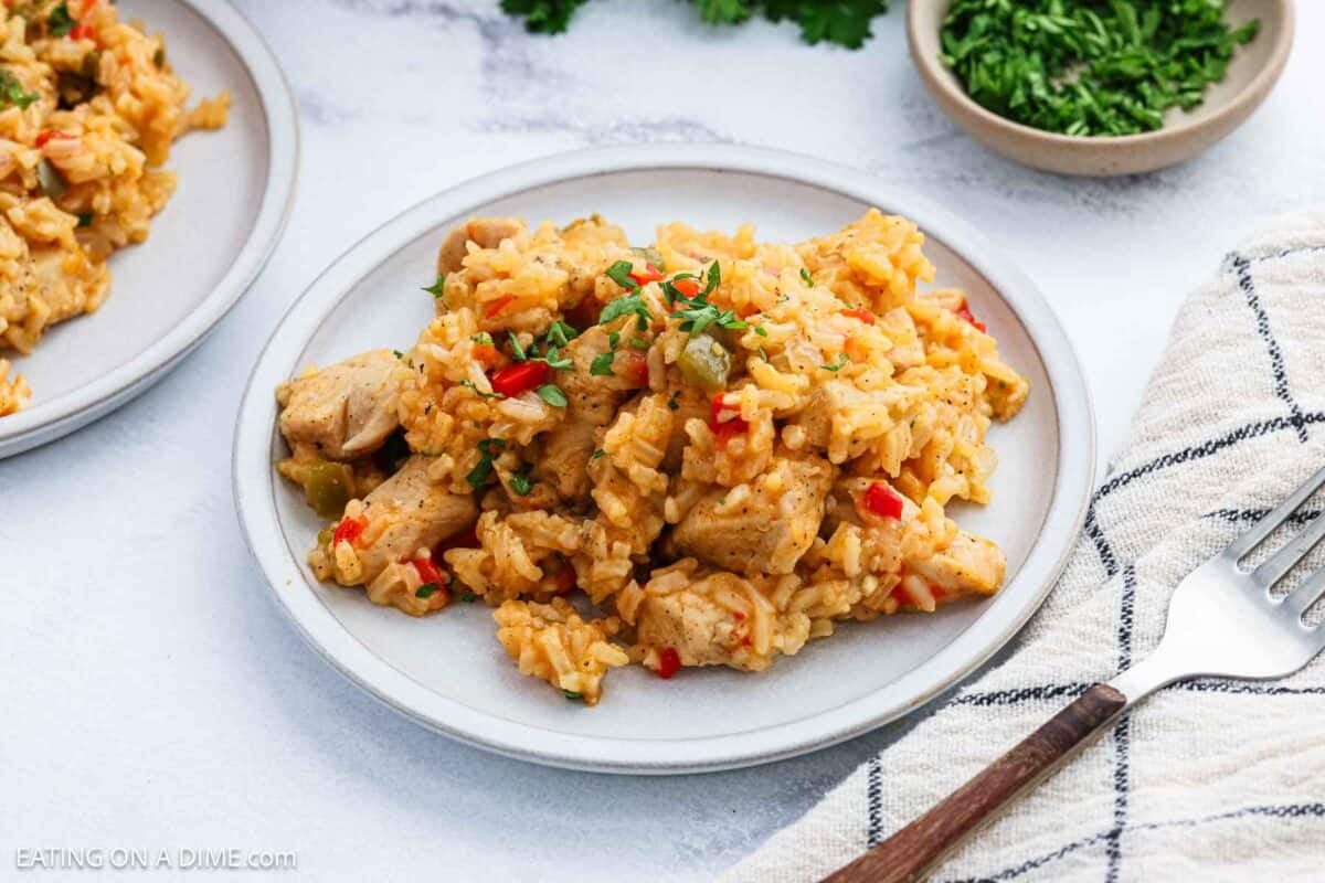 A plate of Cajun chicken and rice with red and green bell peppers, garnished with chopped herbs. A fork rests on a striped napkin nearby, and a small bowl of chopped herbs is in the background.