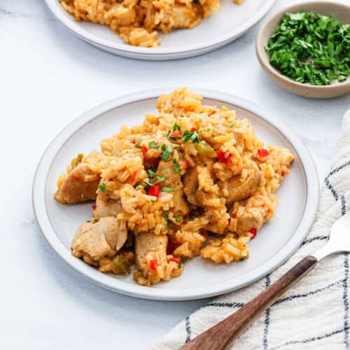 A plate of Cajun Chicken and Rice with diced vegetables, garnished with chopped herbs, sits on a white surface next to a small bowl of fresh parsley and a striped napkin with a fork.