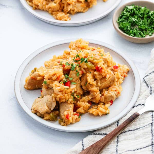 A plate of Cajun Chicken and Rice with diced red and green peppers, garnished with chopped parsley. A small bowl of parsley and a fork on a striped cloth napkin are beside the plate.