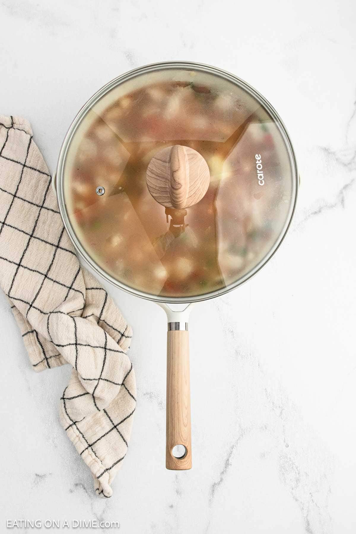 A skillet with a glass lid and wooden handle sits on a marble surface next to a white and black checkered kitchen towel. Inside, delicious Cajun Chicken and Rice is steaming, visible through the fogged lid.
