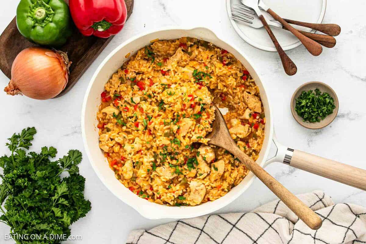 A skillet filled with Cajun Chicken and Rice garnished with herbs sits on a white surface. Surrounding it are fresh parsley, onion, bell peppers, a bowl of chopped herbs, stacked plates, and a set of forks.