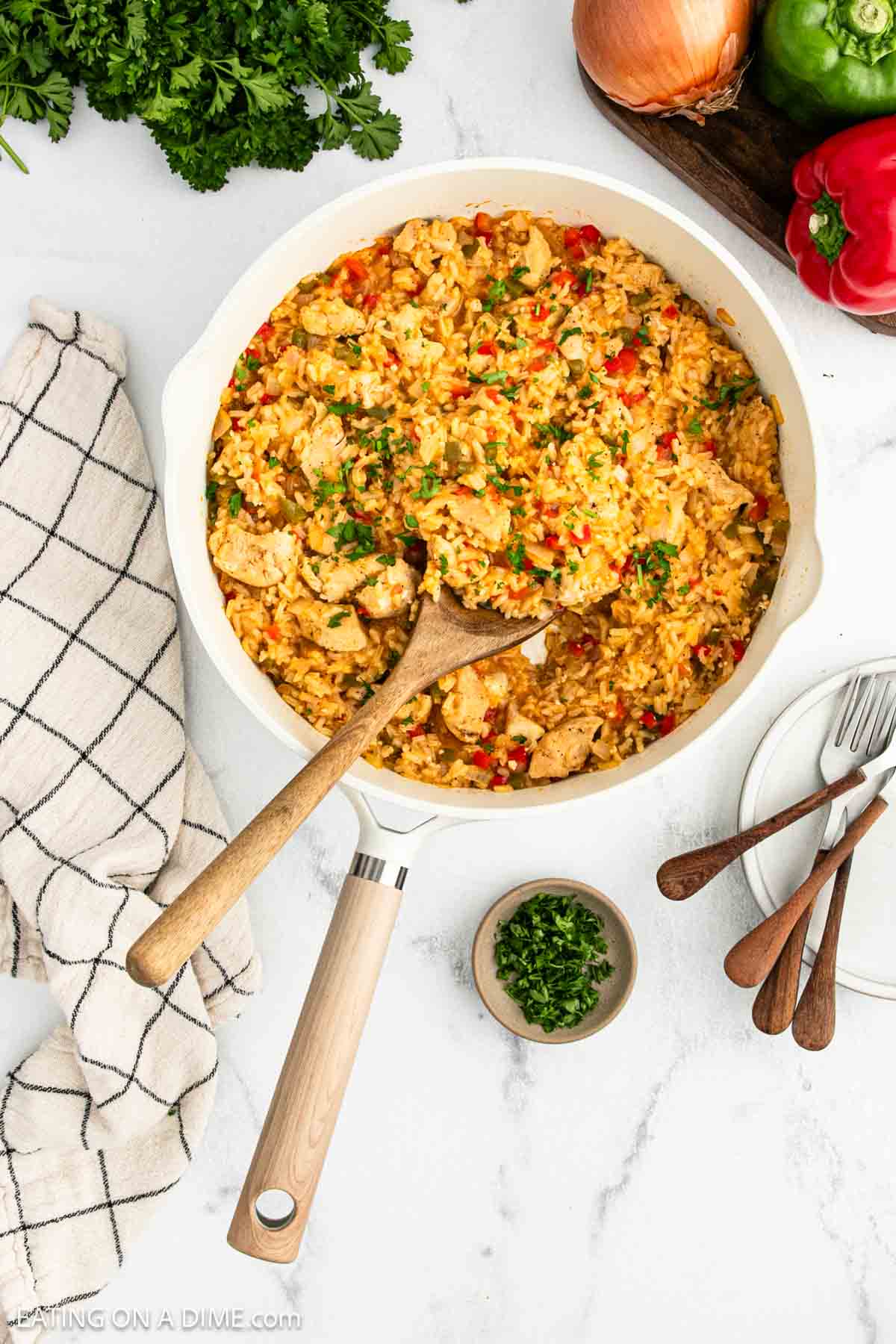 A skillet filled with Cajun Chicken and Rice mixed with red bell peppers and herbs sits on a white surface. A wooden spoon rests in the skillet. Fresh parsley, plates, forks, an onion, and a towel are nearby.