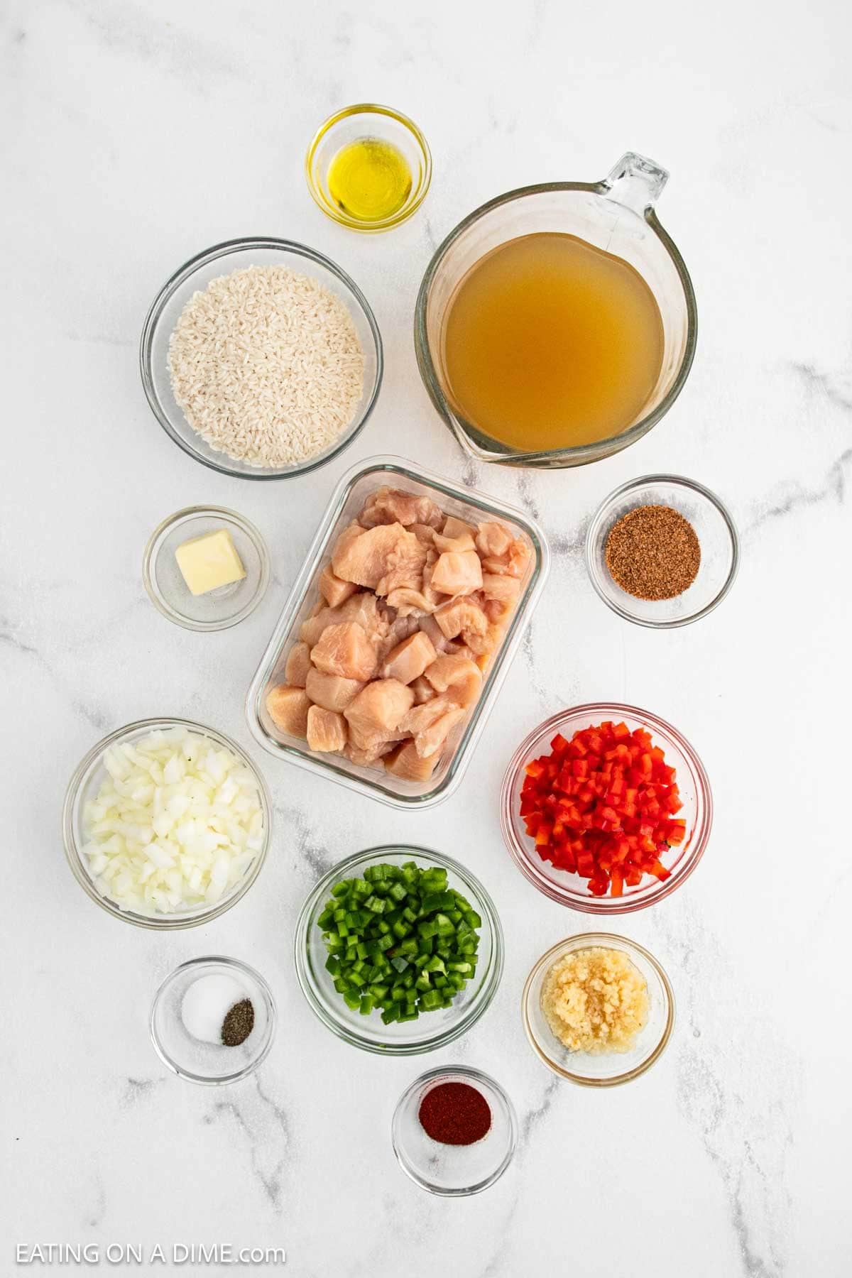 Various ingredients on a white surface, including diced raw Cajun Chicken, rice, chicken broth, chopped onions, bell peppers, minced garlic, butter, olive oil, and small bowls of spices for flavorful Cajun Rice.