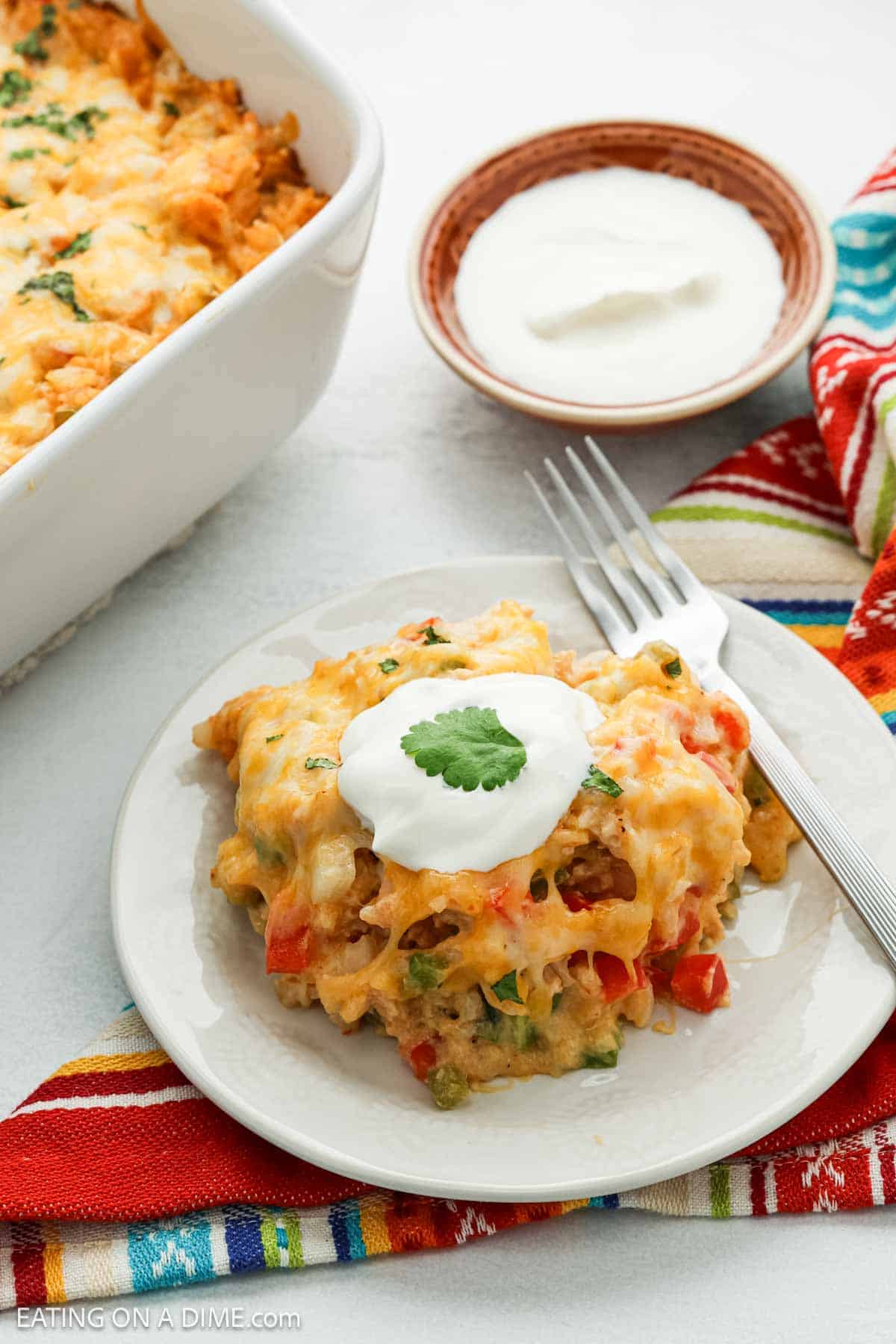 A serving of cheesy Chicken Fajita Casserole with vegetables on a white plate, topped with a dollop of sour cream and a cilantro leaf, next to a fork. In the background are a casserole dish and a bowl of sour cream.