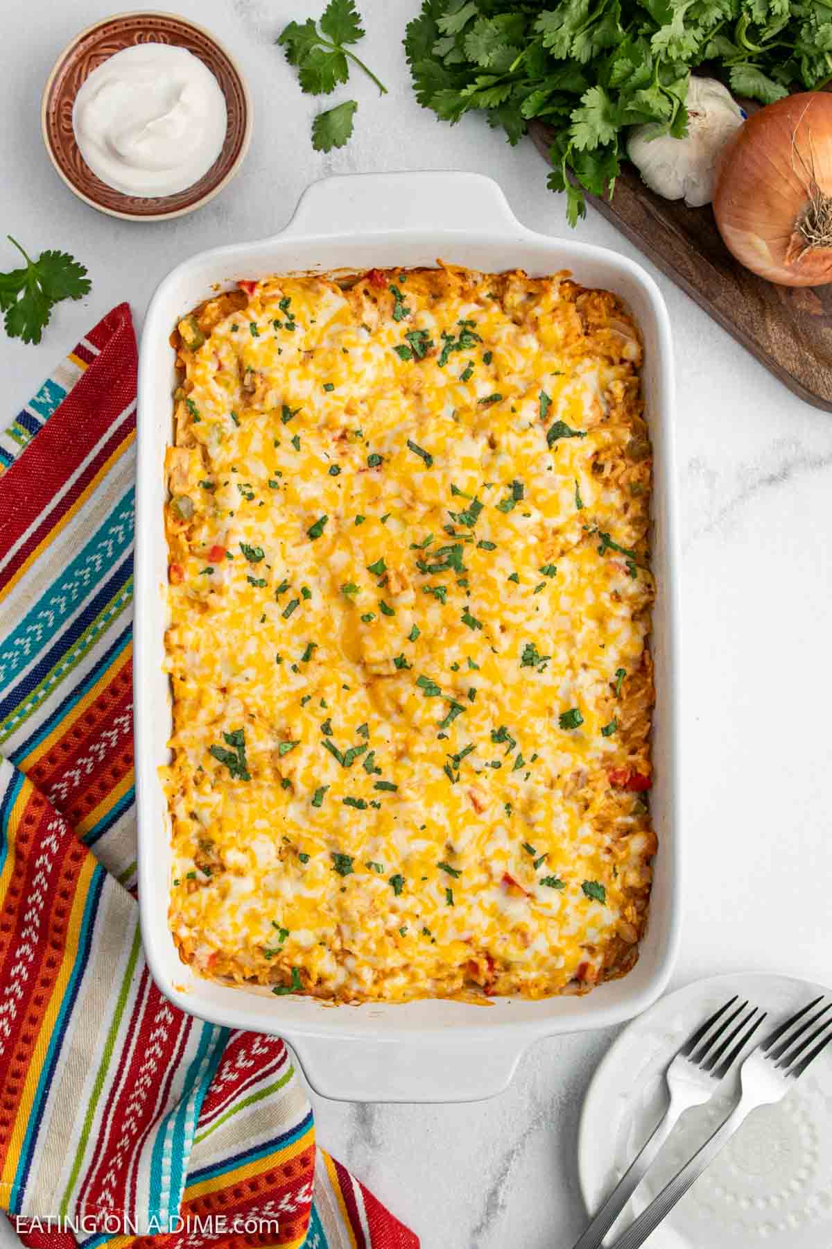 A casserole dish filled with baked cheesy Chicken Fajita Casserole, topped with melted cheese and chopped cilantro. Surrounding the dish are a bowl of sour cream, fresh cilantro, an onion, garlic, and a colorful napkin.