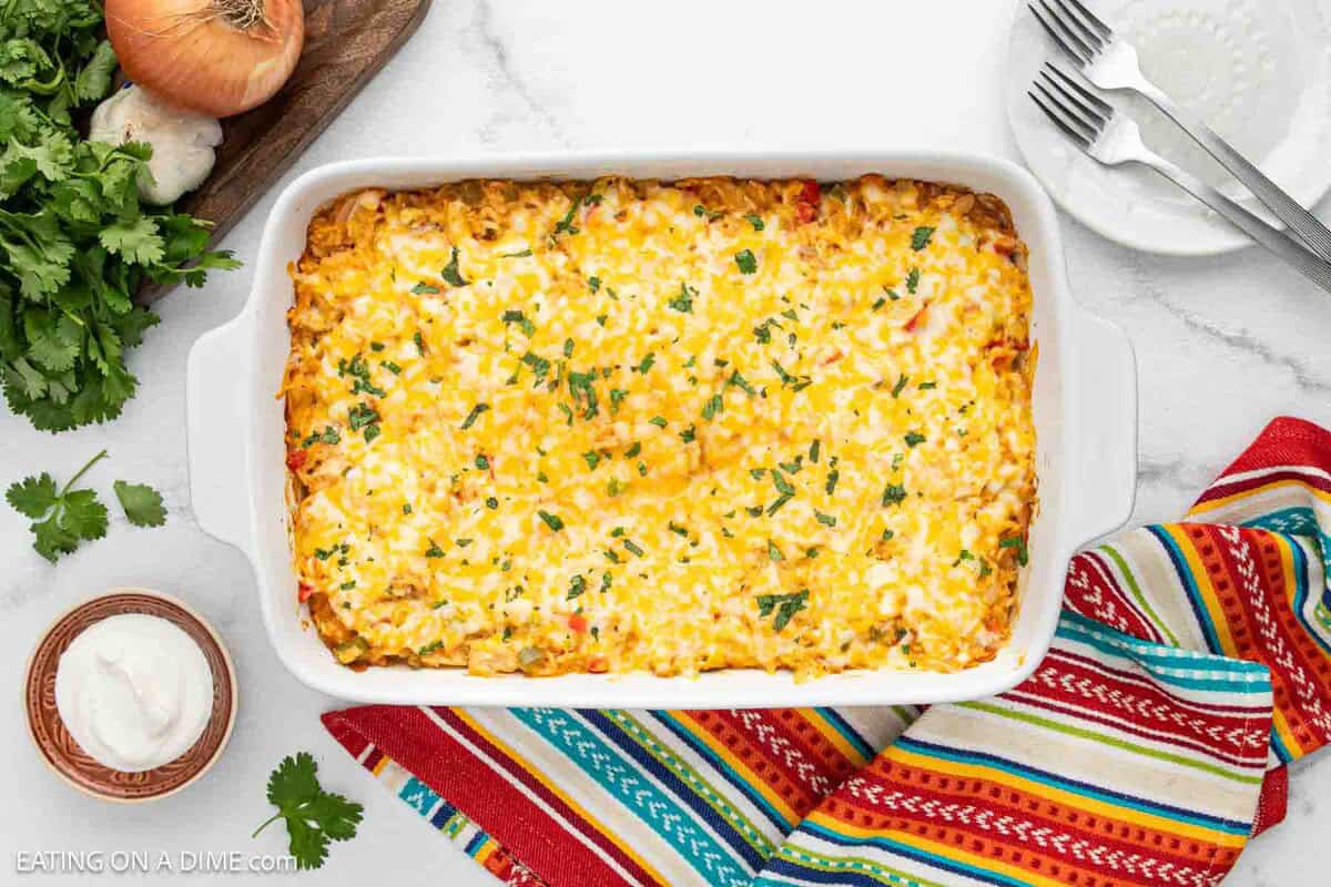 A casserole dish filled with a baked, cheesy Mexican rice Chicken Fajita Casserole garnished with herbs. Surrounding the dish are a colorful striped towel, a small bowl of sour cream, fresh cilantro, an onion, and stacked plates with forks.
