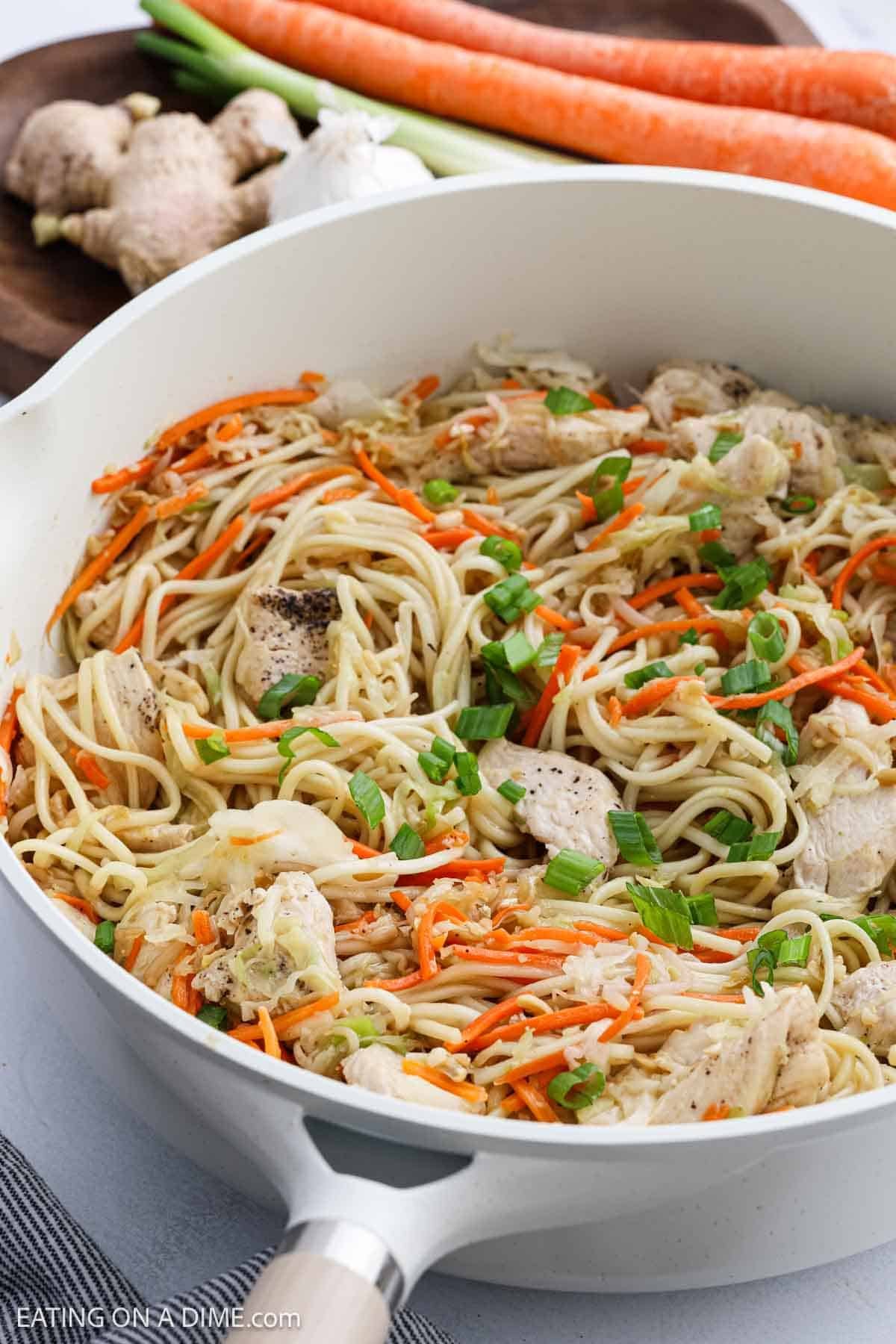 A white skillet filled with cooked noodles, sliced chicken, shredded carrots, and chopped green onions. In the background, fresh ginger, garlic, and whole carrots are visible on a wooden board.