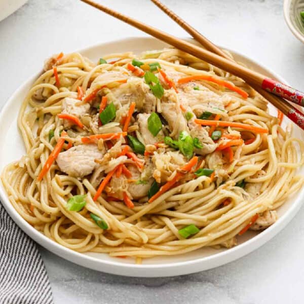 A plate of Chow Mein noodles mixed with shredded chicken, julienned carrots, chopped green onions, and a light sauce. Chopsticks rest on the edge of the plate.