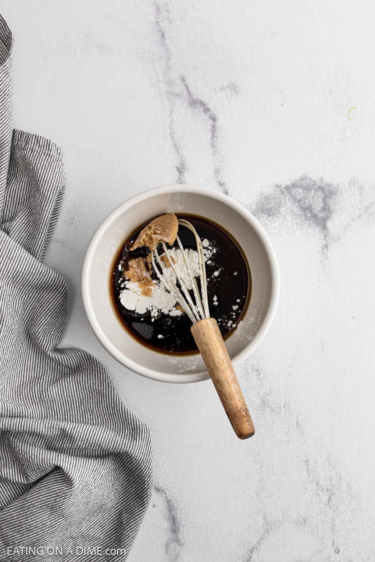 A small white bowl contains dark liquid, white powder, and brown sugar, with a whisk resting inside. The bowl sits on a light surface next to a gray striped cloth.