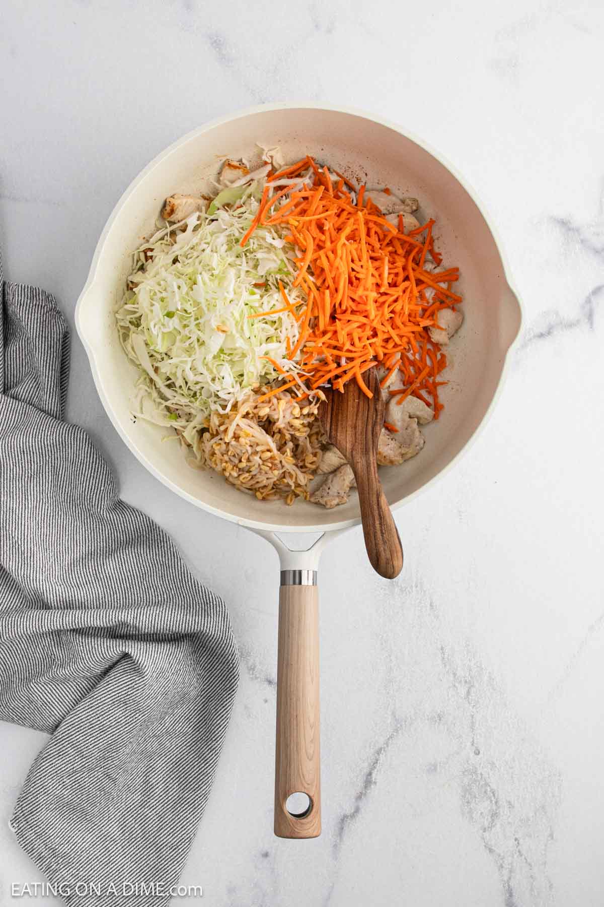 A white skillet on a marble surface contains shredded cabbage, julienned carrots, and pieces of chicken being cooked. A wooden spoon rests inside the skillet, and a gray-striped towel lies beside it.