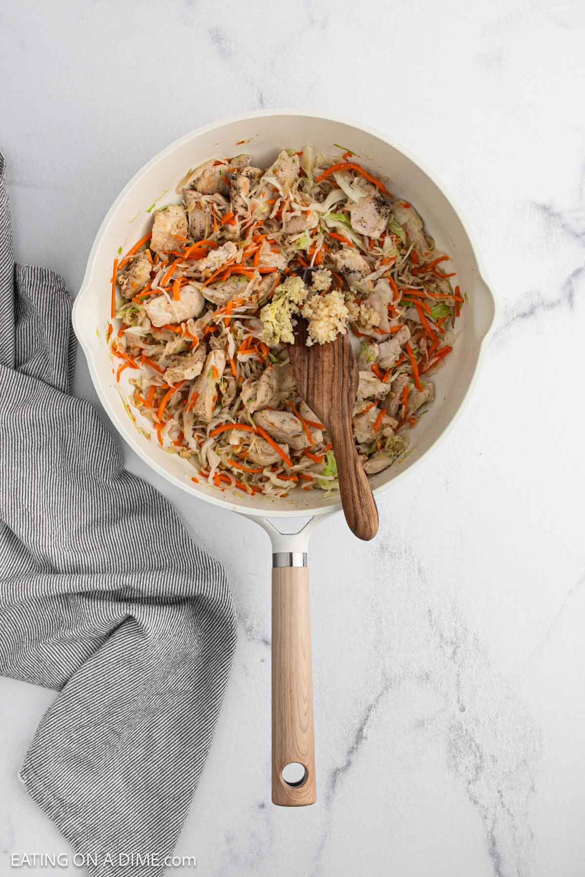 A skillet with cooked shredded chicken, carrots, cabbage, and minced garlic on a wooden spoon, sits on a white marble countertop next to a gray striped towel.