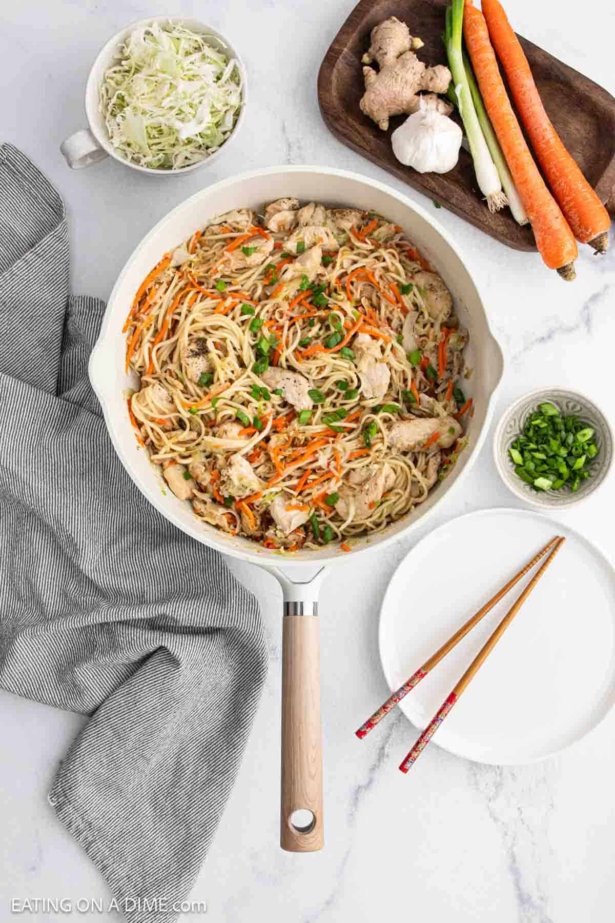 A skillet filled with chicken lo mein, noodles, shredded carrots, and green onions is on a marble counter. Surrounding it are bowls of shredded cabbage, sliced green onions, whole carrots, ginger, garlic, and empty plates with chopsticks.
