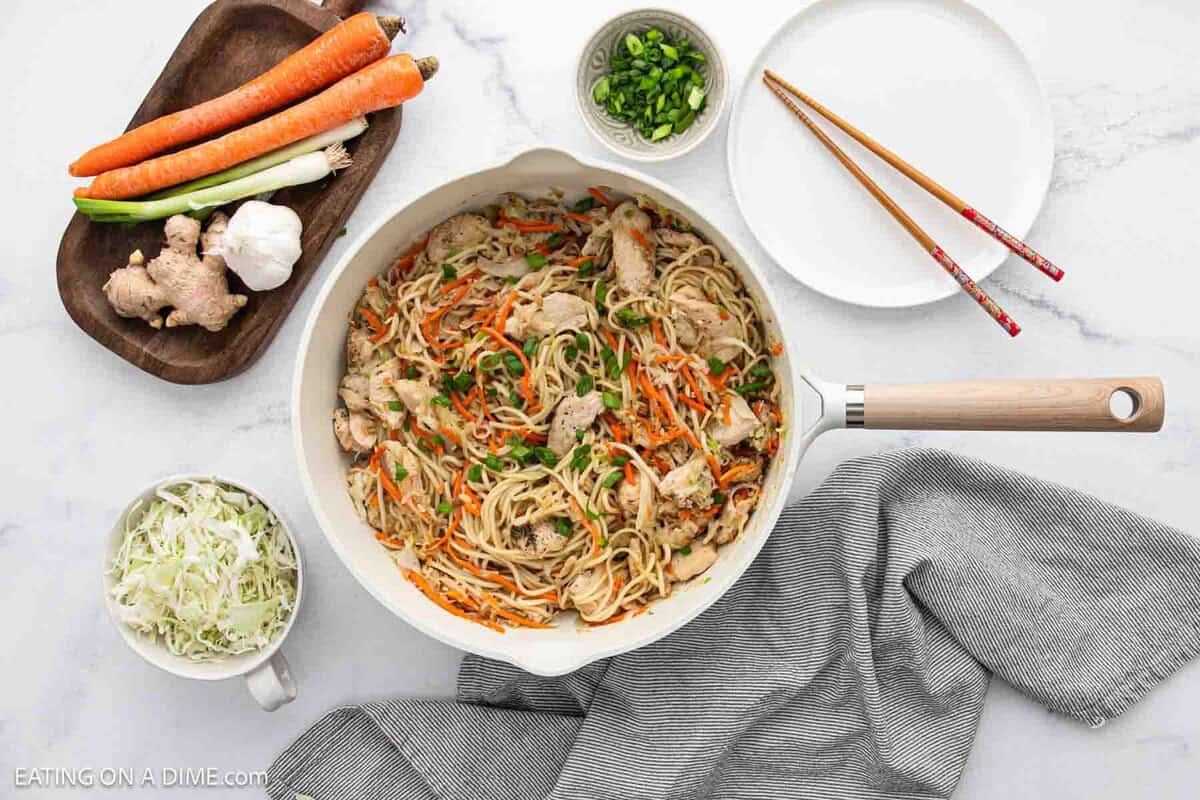 A skillet of lo mein noodles with chicken, shredded carrots, and green onions sits on a marble surface, surrounded by a plate, chopsticks, sliced green onions, a gray towel, and fresh vegetables on a wooden tray.