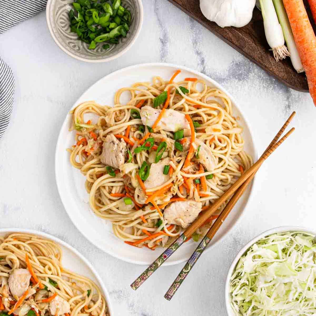 A plate of chicken lo mein noodles with sliced carrots and green onions, served with chopsticks. Nearby are bowls of green onions, shredded cabbage, and whole vegetables on a light-colored table.