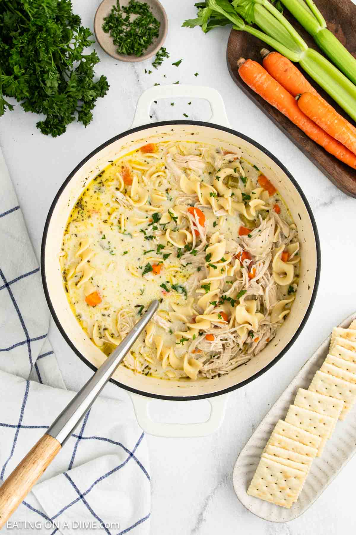 A pot of creamy chicken noodle soup with shredded chicken, carrots, celery, and egg noodles, garnished with chopped herbs. Crackers, fresh vegetables, and parsley surround the creamy chicken noodle soup on a white surface.
