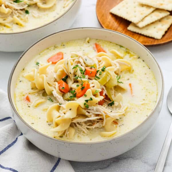 A bowl of Creamy Chicken Noodle Soup with egg noodles, shredded chicken, carrots, and celery, garnished with parsley. Crackers rest on a plate in the background while a spoon and napkin sit beside the bowl.