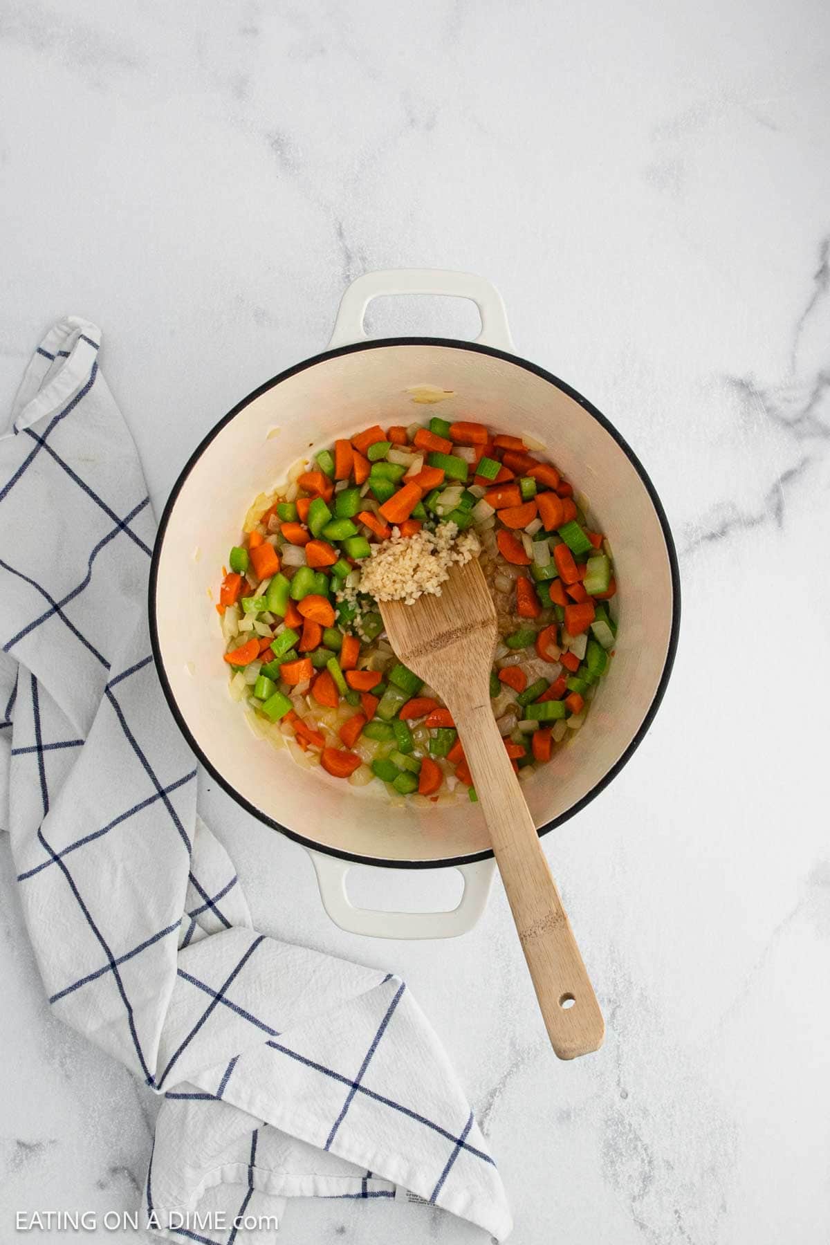 A white pot for Creamy Chicken Noodle Soup is filled with chopped onions, carrots, celery, and minced garlic on a white countertop next to a blue-striped dish towel. A wooden spoon rests in the pot.