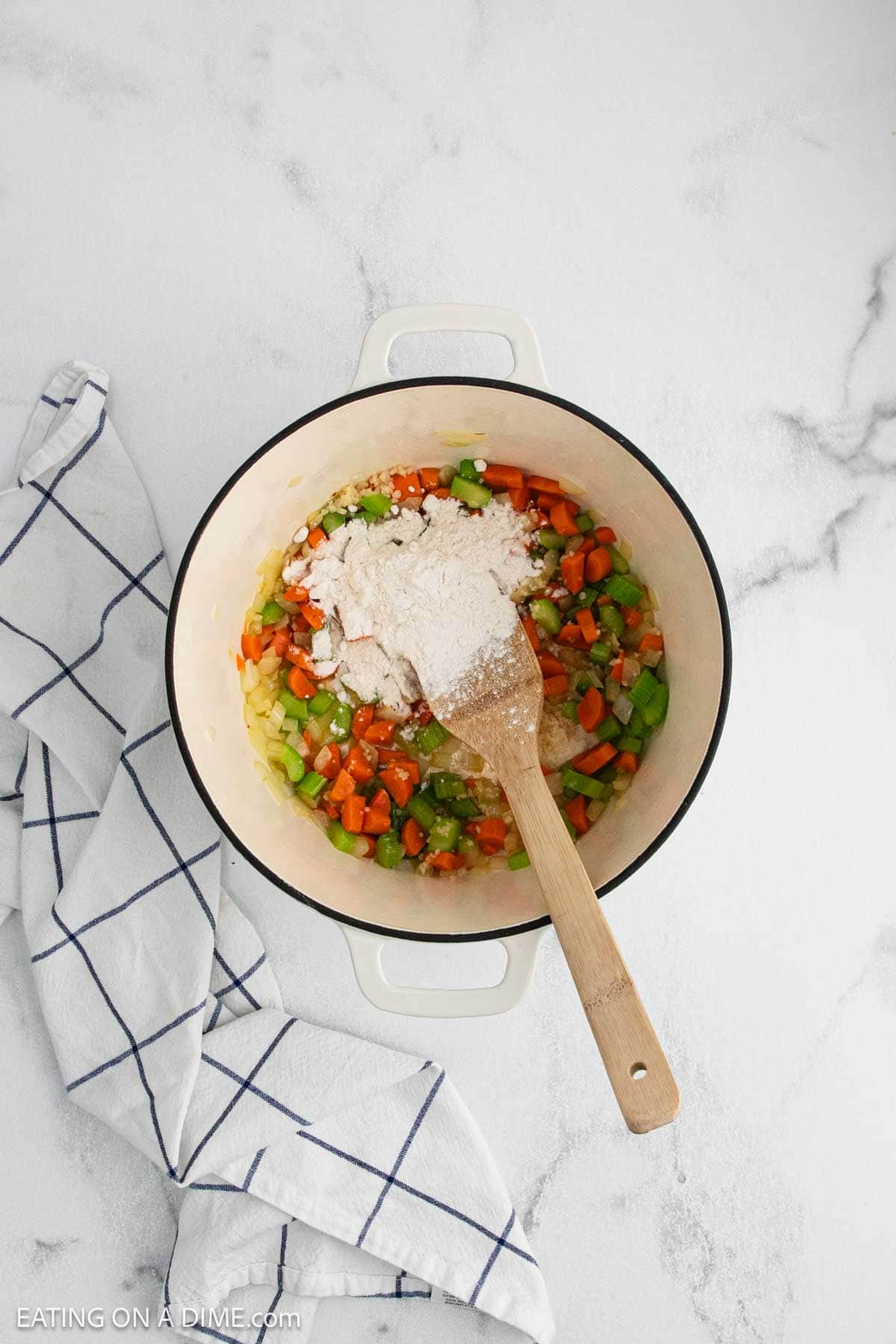 A white pot filled with chopped onions, carrots, celery, and flour—key ingredients for Creamy Chicken Noodle Soup—sits on a white marble surface next to a striped towel. A wooden spoon rests inside the pot.