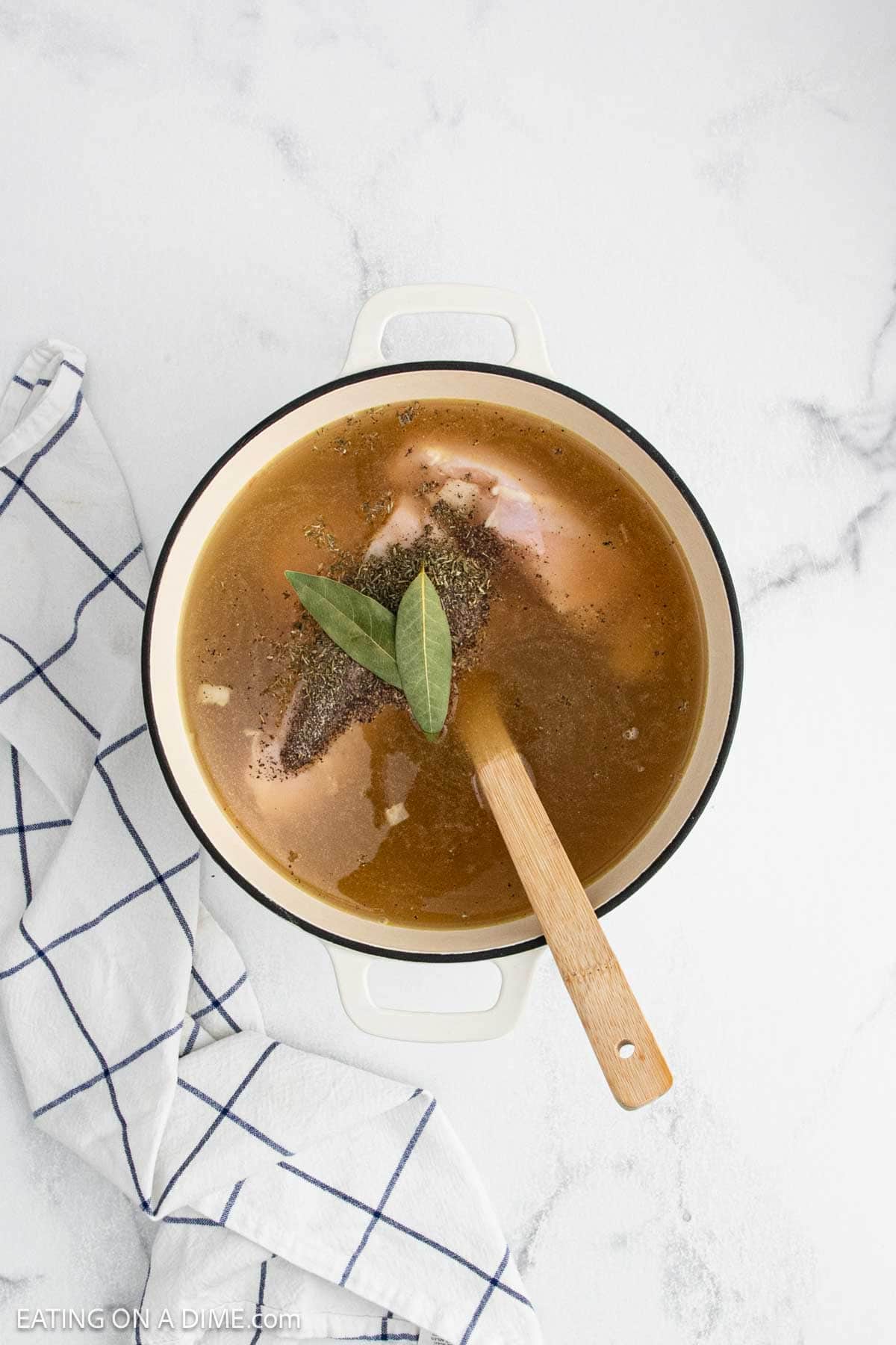 A white pot filled with creamy chicken noodle soup, broth, spices, and two bay leaves sits on a white surface next to a white kitchen towel with blue grid lines. A wooden spoon rests in the pot.