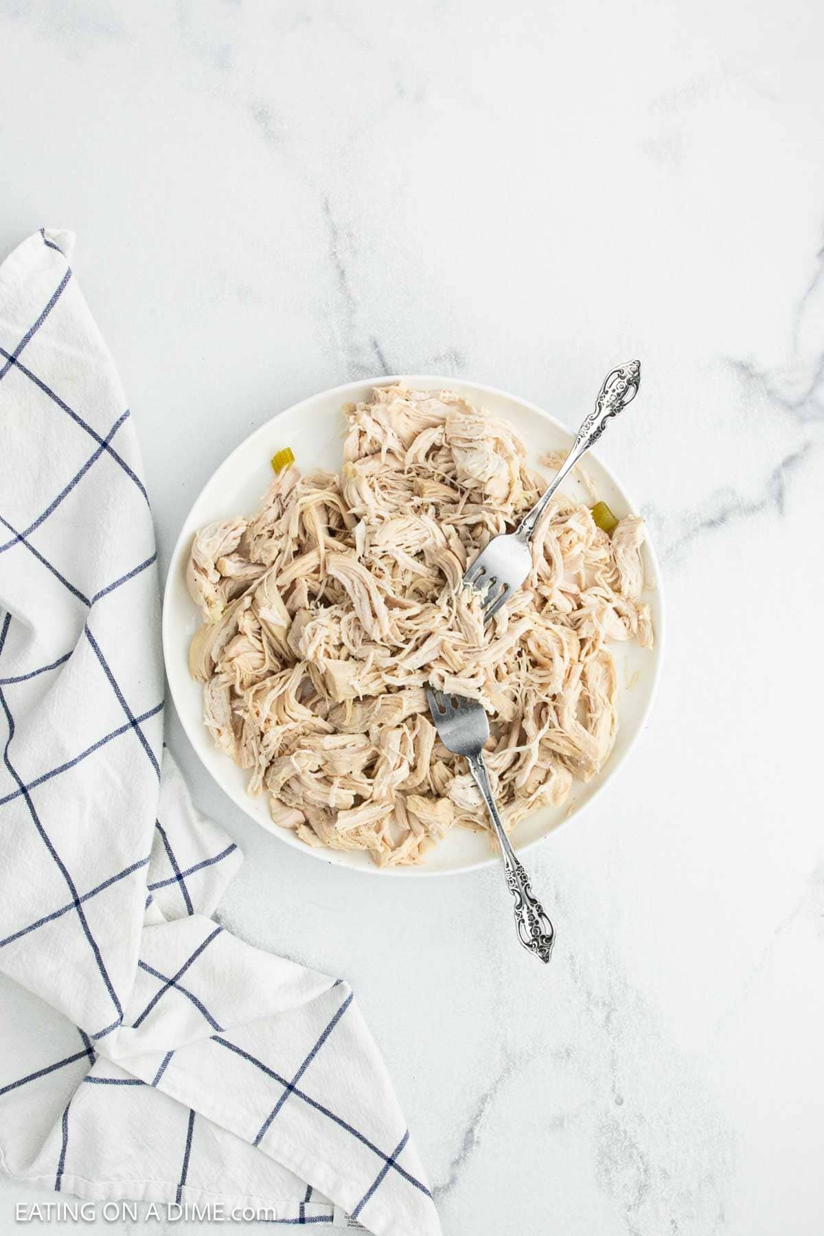 A plate of shredded chicken with two forks on a white surface, next to a white and blue checkered kitchen towel—perfect for making creamy chicken noodle soup.