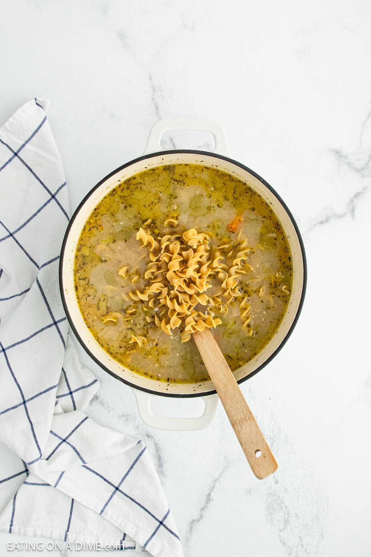 A white pot filled with creamy chicken noodle soup and uncooked spiral egg noodles sits on a white marble surface, with a blue and white checkered kitchen towel beside it. A wooden spoon rests in the pot.