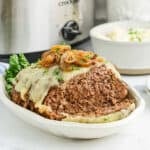 A sliced French Onion Meatloaf topped with melted cheese and caramelized onions sits on a white serving dish, garnished with parsley. A crockpot and a bowl of mashed potatoes are in the background.