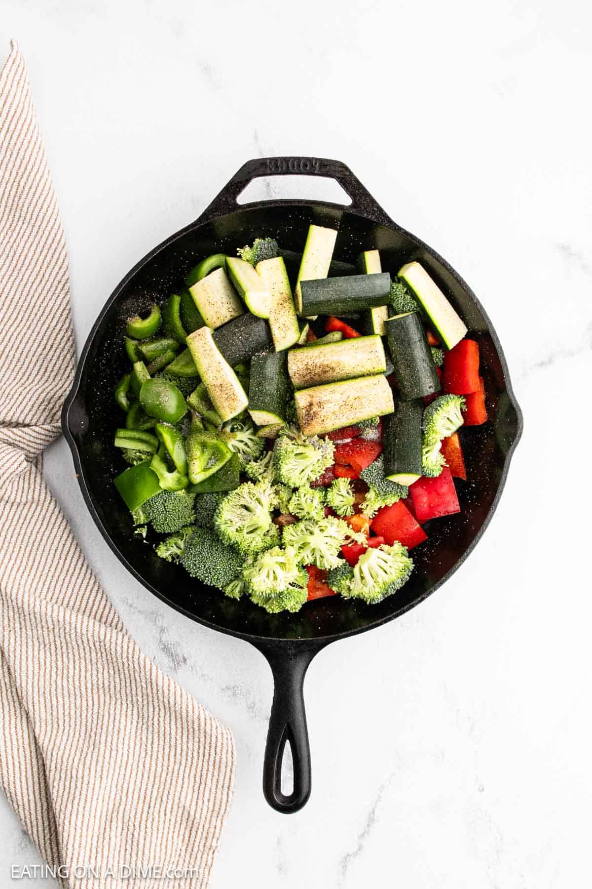 A cast iron skillet filled with chopped broccoli, red bell pepper, green bell pepper, and zucchini—classic Hibachi Vegetables—topped with seasoning, sits on a white surface next to a striped kitchen towel.