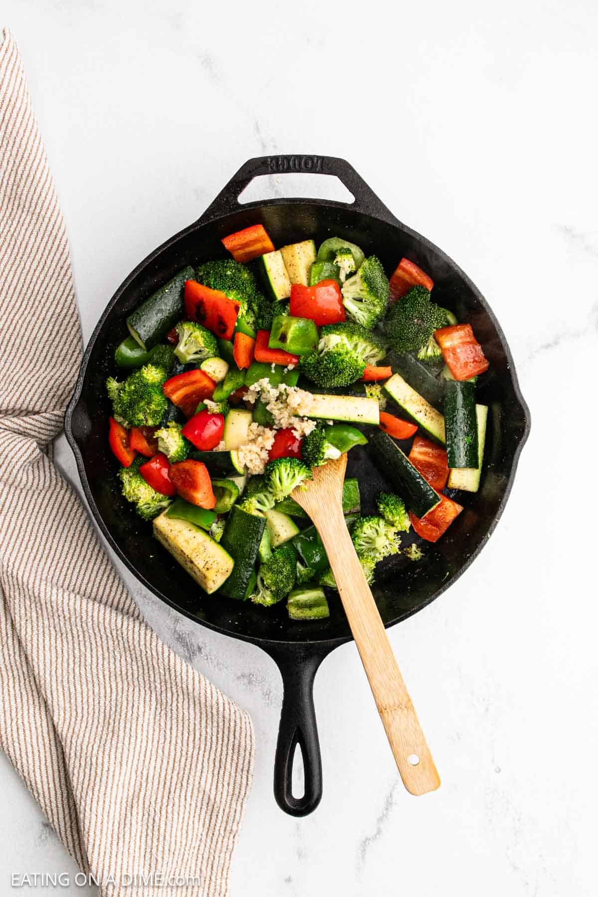 A cast iron skillet filled with colorful chopped hibachi vegetables—broccoli, zucchini, green bell pepper, and red bell pepper—sits on a white surface next to a striped kitchen towel. A wooden spoon rests in the skillet.