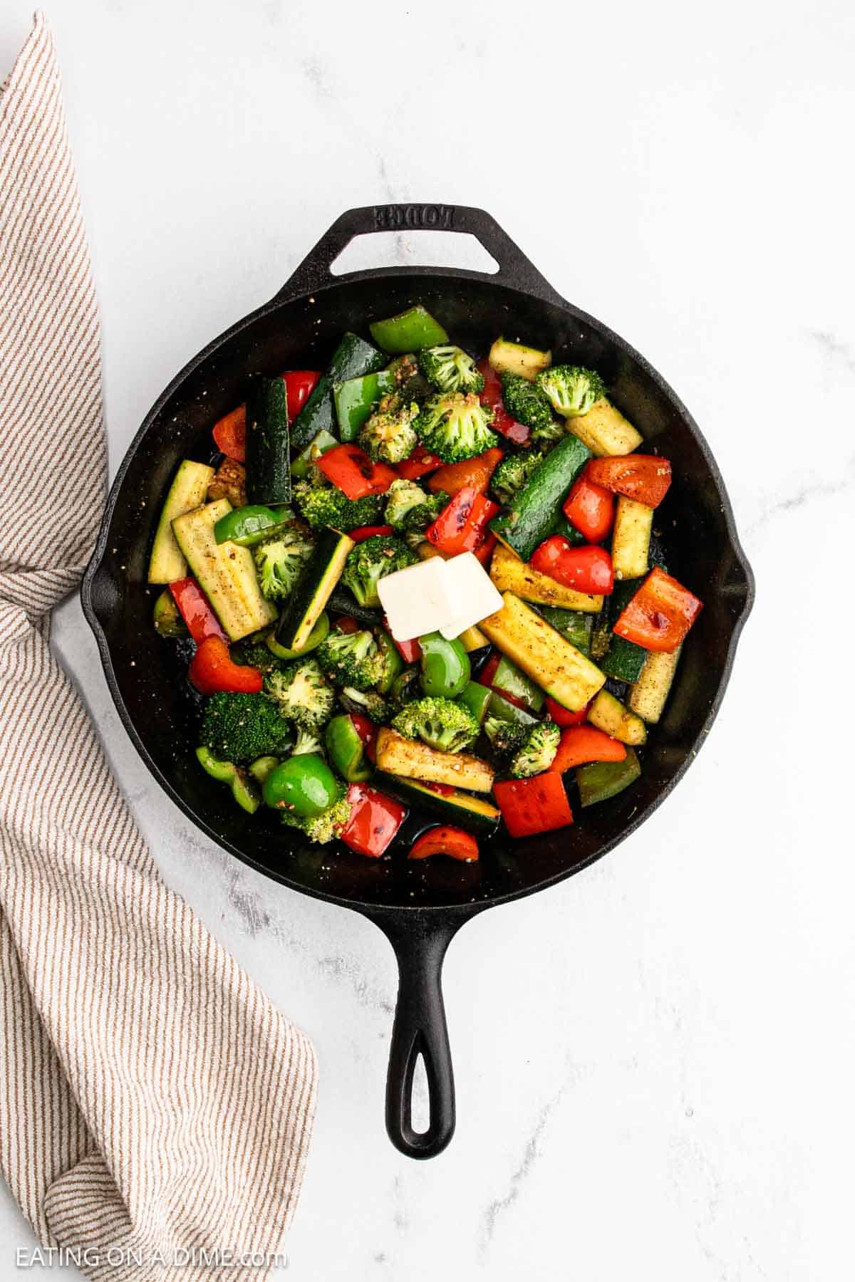 A cast iron skillet filled with colorful hibachi vegetables, including broccoli, zucchini, bell peppers, and snap peas, topped with two cubes of butter. A striped kitchen towel is nearby on a white surface.