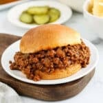 A Sloppy Joes sandwich filled with seasoned ground beef on a white plate, served with sliced pickles and a bowl of chips in the background—perfect for fans of classic Manwich flavor.