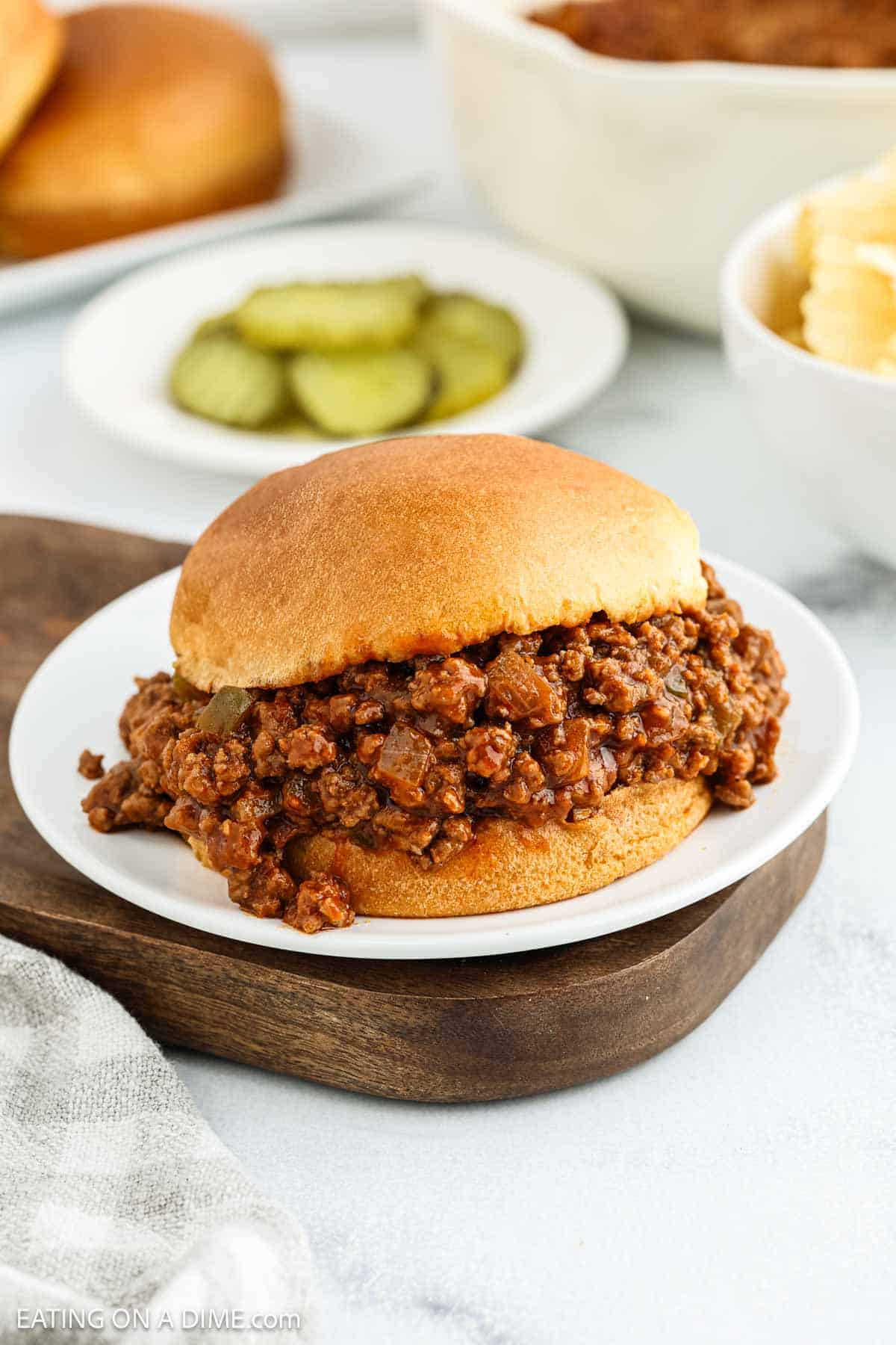 A classic Sloppy Joes sandwich filled with seasoned ground beef sits on a white plate. In the background, there are pickle slices, potato chips, and part of a bun visible.
